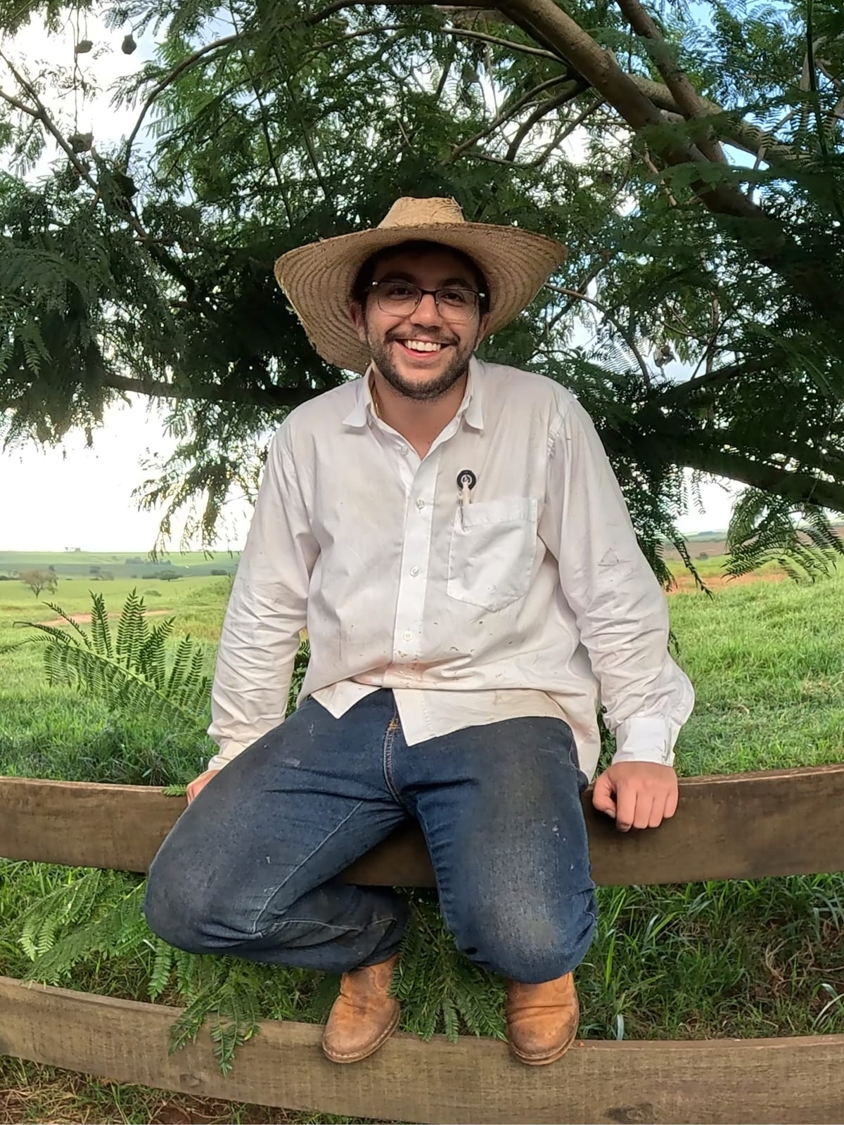Pedro Henrique, de chapéu, sentado na cerca da fazenda com o pasto ao fundo